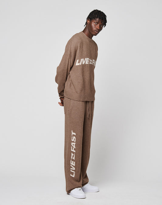 A person poses against a plain background in an oversize fit Live Fast Boucle Sweater by LIVE FAST DIE YOUNG and matching pants, both brown with LIVEFAST in white, styled with white sneakers.