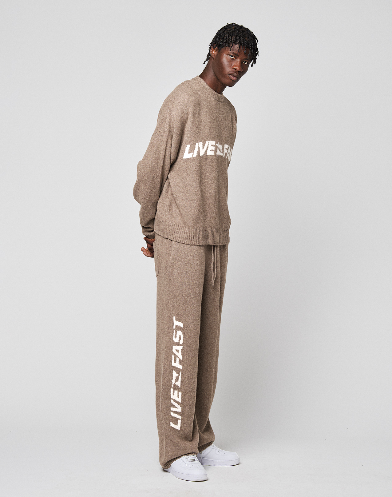 A person wears the LIVE FAST DIE YOUNG Live Fast Boucle Sweater and matching beige pants, both with “LIVE FAST” in white text and an oversize fit, standing against a plain white background and looking slightly to the side.