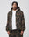 A man with braided hair wears a LIVE FAST DIE YOUNG LFDY Fleece Hooded Zip in an oversize camouflage pattern, paired with matching pants and a white t-shirt, standing against a plain light gray background.