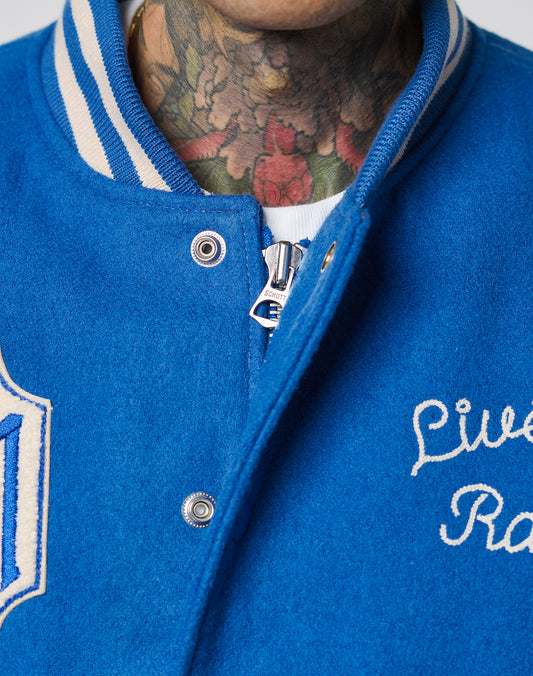 Close-up of a person in a LIVE FAST DIE YOUNG LFDY x Schott NYC Varsity Jacket with chenille patches, cowhide leather sleeves, white trim, and embroidered text. Colorful floral neck tattoos are visible above a white shirt.