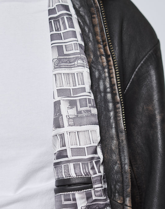 A close-up of a person in a white shirt and a black LIVE FAST DIE YOUNG LFDY Leather Jacket featuring an inner lining with a black-and-white Plattenbau Print building facade pattern.
