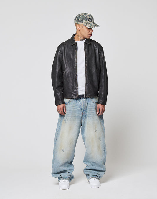 Wearing a camouflage cap, white t-shirt, very baggy faded jeans, white sneakers, and a LIVE FAST DIE YOUNG LFDY Leather Jacket, a person stands against a plain white background, looking slightly to their left.