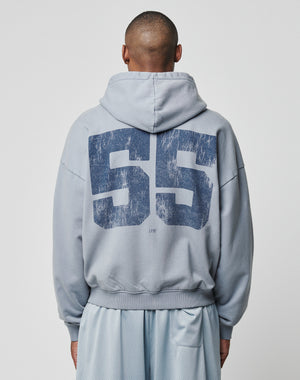 A person faces away from the camera wearing the LFDY 55 Hooded Zip by LIVE FAST DIE YOUNG in light gray, featuring a large distressed dark blue “55” in Vintage Waschung on the back. The hood is up and it’s styled with light mesh shorts.