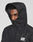 A person wears the LIVE FAST DIE YOUNG Live Fast Performance Rain Jacket in black with the hood up, partially covering their face. This regular fit jacket has a white LIVE FAST logo on the chest and elastic cuffs. The background is plain light gray.