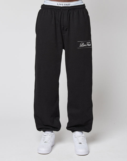 A person wearing black Wide Cuff Jogger sweatpants by LIVE FAST DIE YOUNG, featuring a Live Fast logo on the left thigh, a comfortable elastic waistband, soft French Terry fabric, and paired with white sneakers against a plain white background.