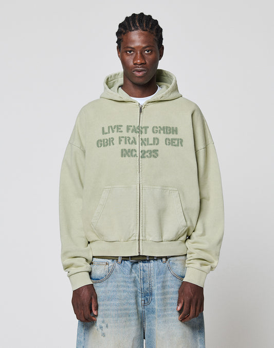 A person stands in light blue jeans against a plain background, wearing a LIVE FAST DIE YOUNG LFDY District Hooded Zip—an oversize fit light green zip-up hoodie made from 100% cotton, with "LIVE FAST GMBH GBR FRA.GLD GER INC. 235" on the front.