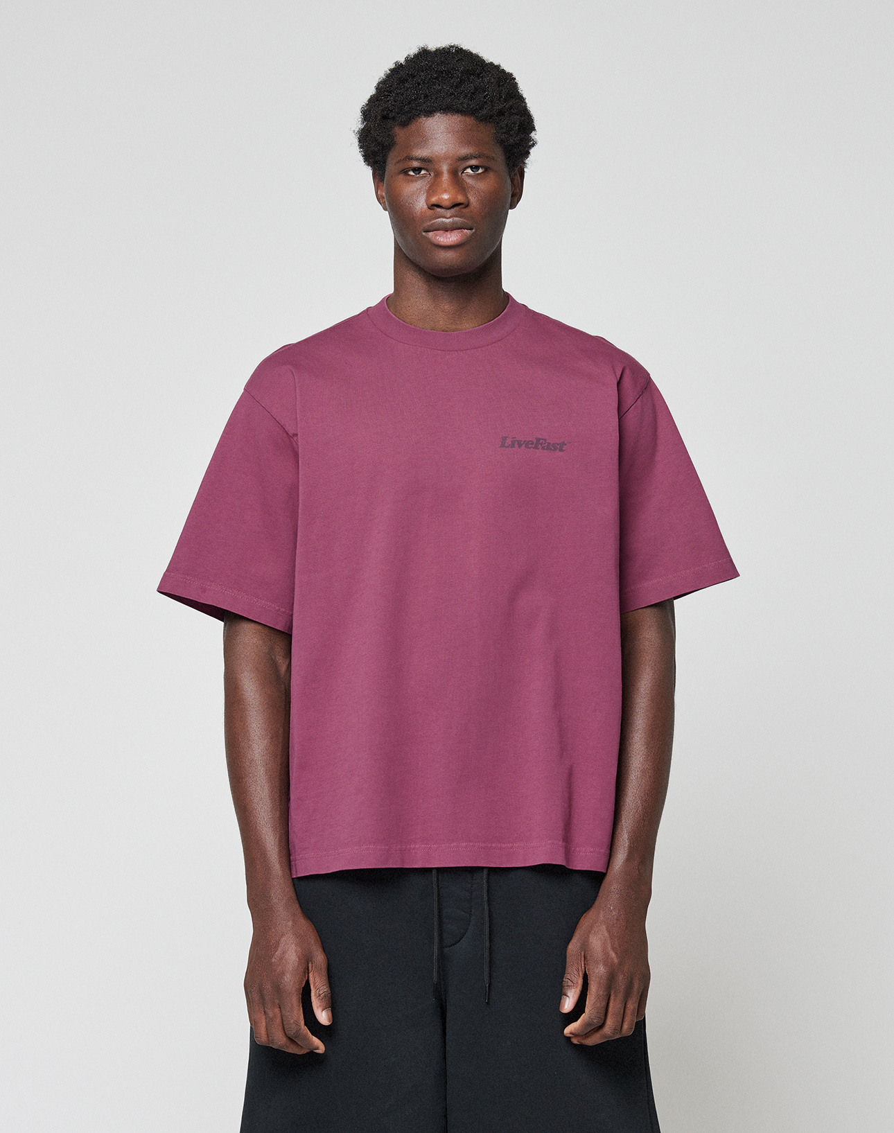A person faces forward in a loose-fitting maroon Basic 210 Tee by LIVE FAST DIE YOUNG, featuring "LiveFast" on the chest, made from 100% cotton, paired with black shorts against a plain light gray background.