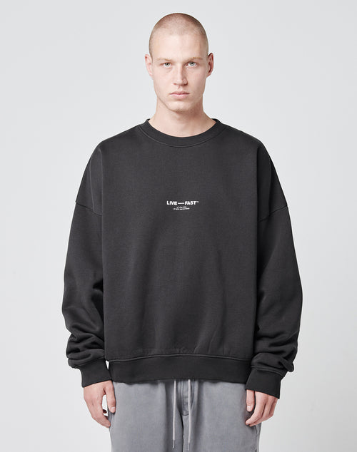 A person with a shaved head models the Tape Sweater by LIVE FAST DIE YOUNG—an oversized black sweatshirt with LIVE FAST in small white text on the chest—paired with light gray pants against a plain light gray background.