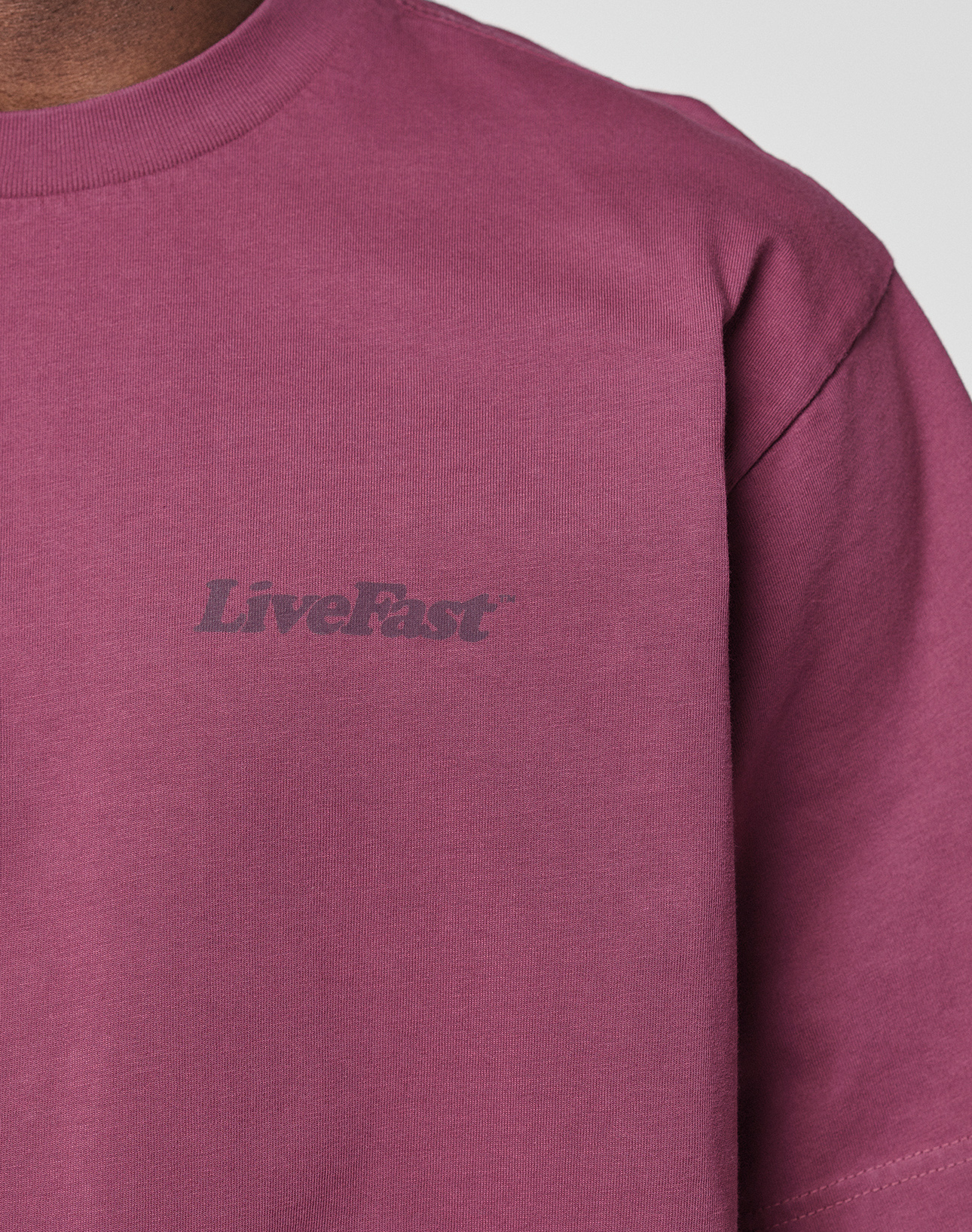 Close-up of a person wearing the LIVE FAST DIE YOUNG Basic 210 Tee, a maroon 100% Baumwolle T-shirt with "LiveFast" in a darker shade on the left chest. Only the upper chest, shoulder, and neck are shown.