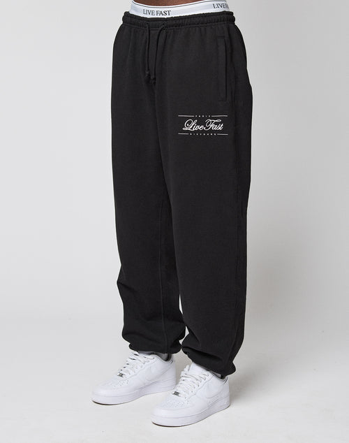 A person wears black Wide Cuff Joggers by LIVE FAST DIE YOUNG, featuring "Live Fast" on the left thigh, elastic waistband, and cuff. Made from soft French Terry. Styled with white sneakers against a plain light background.