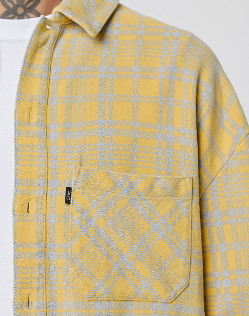 Close-up of a person in the LIVE FAST DIE YOUNG Vintage Flannel—an oversize yellow and gray plaid 100% Baumwolle shirt with front pocket, black tag, worn over a white tee. Part of a neck tattoo is also visible.