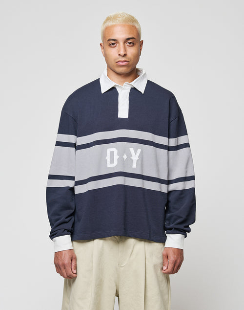 A person with short bleached blonde hair wears the LIVE FAST DIE YOUNG LFDY Striped Polo, an oversized navy and gray long-sleeve shirt with white collar and cuffs, D Y on the chest, and loose beige cotton-twill pants, against a plain background.