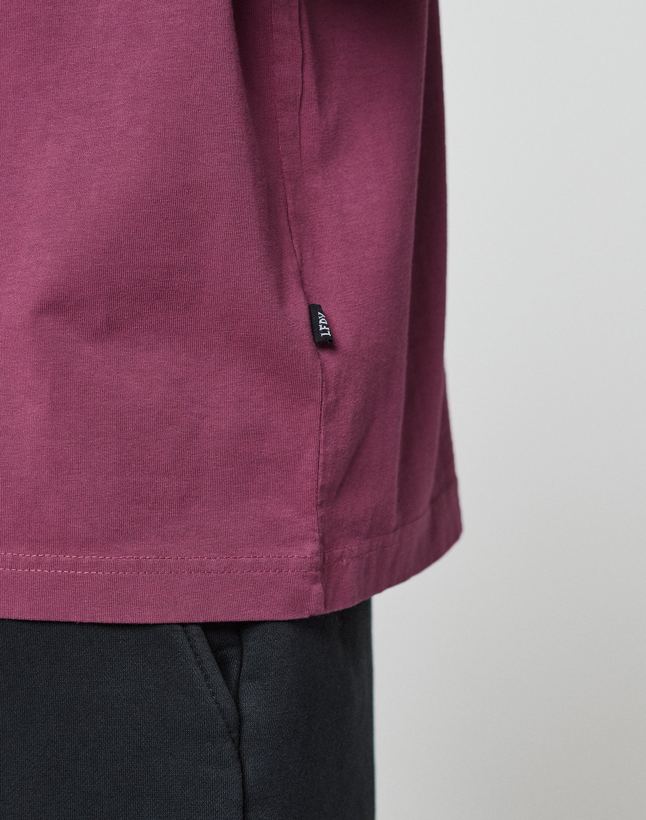 Close-up of a person wearing the LIVE FAST DIE YOUNG Basic 210 Tee in maroon, featuring an oversized fit and a small black tag on the side seam, paired with black pants. The background is plain and light-colored.