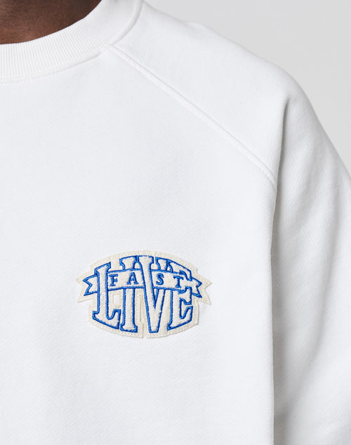 Close-up of the LFDY Radiator Sweater by LIVE FAST DIE YOUNG, an oversized white sweatshirt featuring a bold blue and white chenille FAST LIVE patch on the chest. Only part of the wearers neck and shoulder is shown.