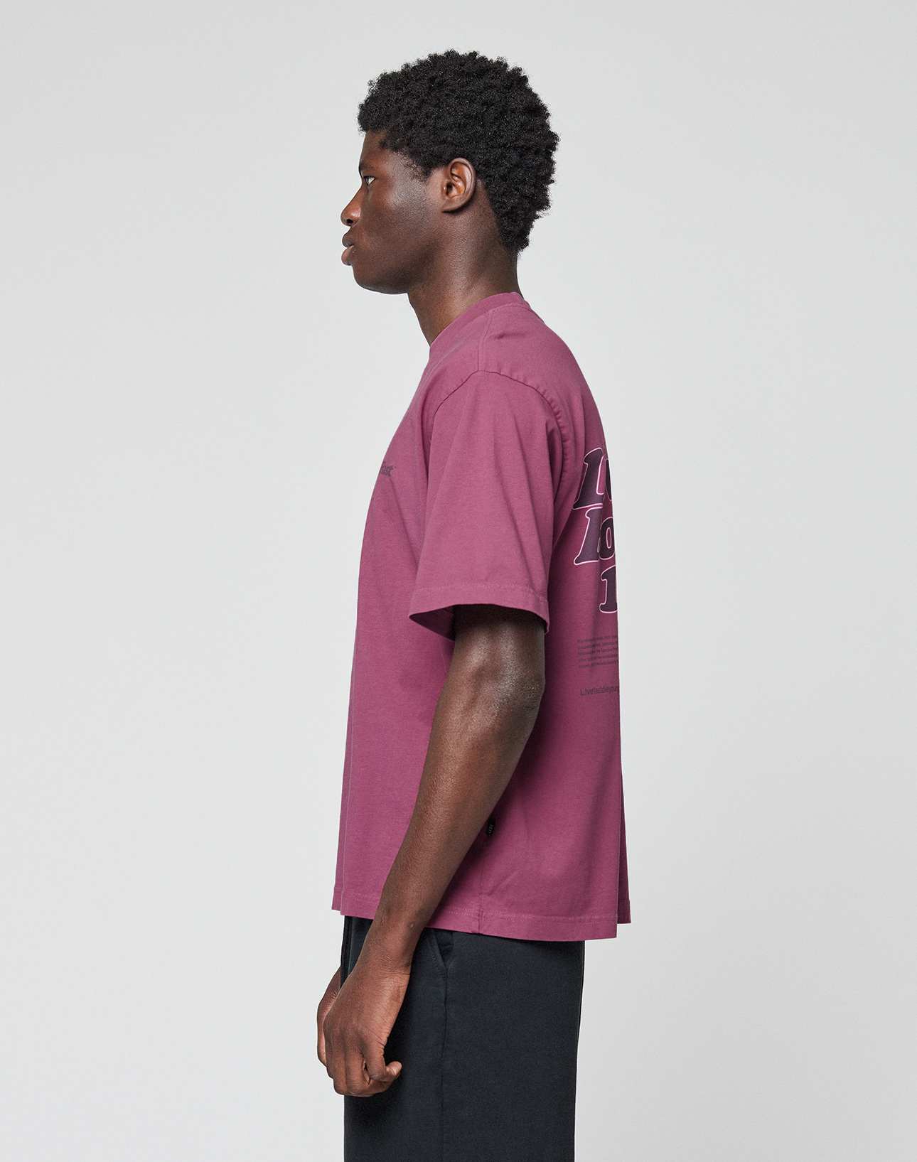 A person stands in profile against a plain background, wearing the LIVE FAST DIE YOUNG Basic 210 Tee in maroon (100% Baumwolle, oversize fit) with black text on the back, paired with black pants. The focus is on their left side.