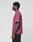 A person stands in profile against a plain background, wearing the LIVE FAST DIE YOUNG Basic 210 Tee in maroon (100% Baumwolle, oversize fit) with black text on the back, paired with black pants. The focus is on their left side.