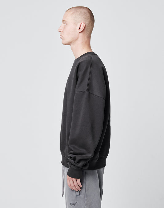 A person with a buzz cut wears the LIVE FAST DIE YOUNG Tape Sweater, an oversize fit dark sweatshirt with cuffed sleeves and hem, paired with light gray pants, standing in profile against a plain light gray background.