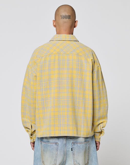 A person with a shaved head and neck tattoo wears a LIVE FAST DIE YOUNG Vintage Flannel—an oversized yellow and gray plaid jacket (100% Baumwolle)—with light blue jeans, facing away from the camera against a plain background.
