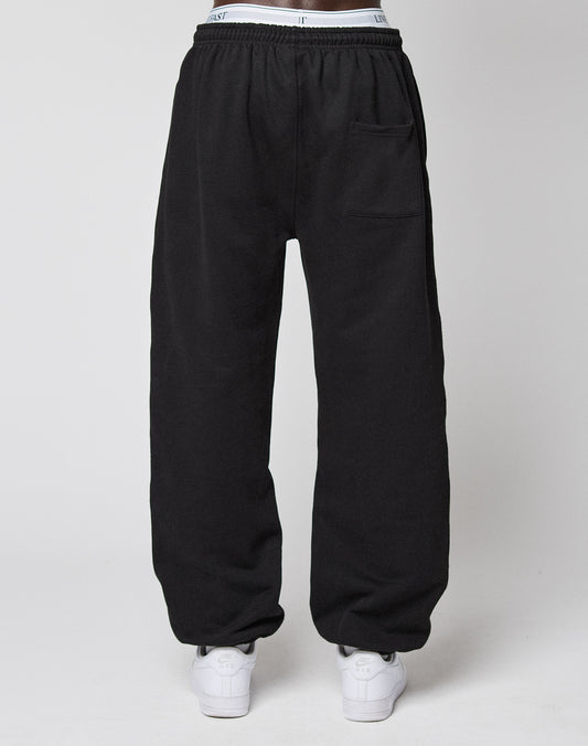 A person wearing LIVE FAST DIE YOUNG Wide Cuff Jogger in black, featuring an elastic waistband and a right thigh pocket, paired with white sneakers, stands against a plain white background.