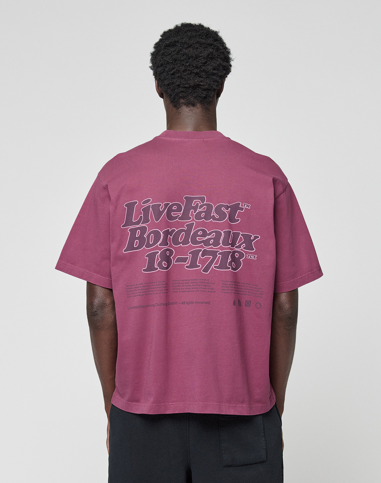 Seen from behind, a person with short curly hair wears the LIVE FAST DIE YOUNG Basic 210 Tee, an oversized maroon 100% Baumwolle shirt with bold “Live Fast Bordeaux 18-1718” front and back prints, paired with black pants.