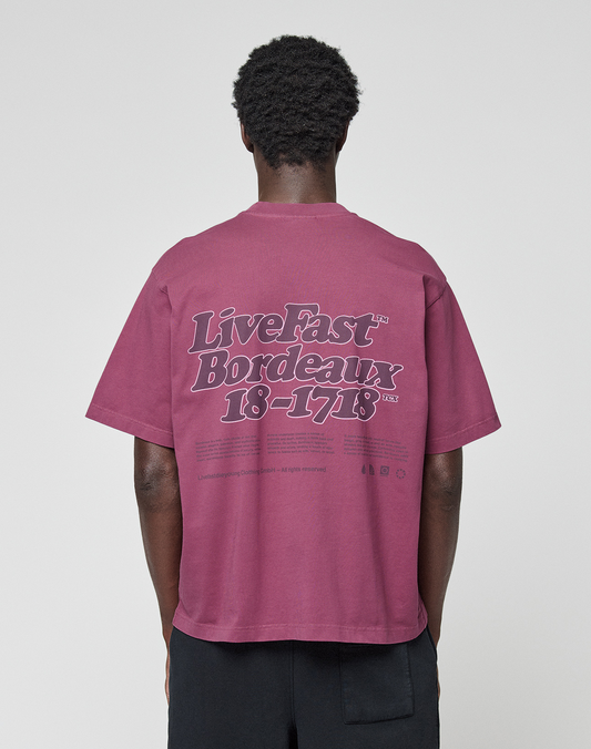 Seen from behind, a person with short curly hair wears the LIVE FAST DIE YOUNG Basic 210 Tee, an oversized maroon 100% Baumwolle shirt with bold “Live Fast Bordeaux 18-1718” front and back prints, paired with black pants.