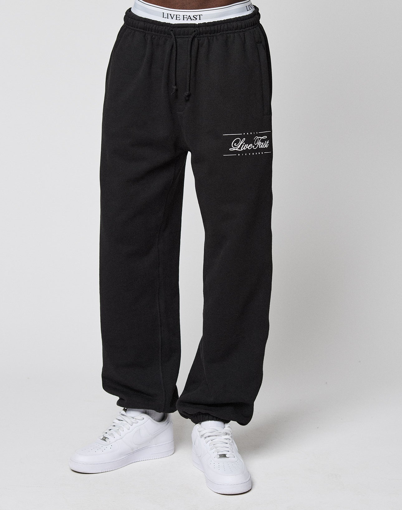A person wears black Wide Cuff Joggers by LIVE FAST DIE YOUNG with a visible French Terry waistband and "LIVE FAST" text, paired with white sneakers, standing against a plain white background.