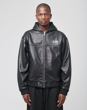 A person faces forward wearing the LIVE FAST DIE YOUNG Leather Hooded Jacket—an oversize fit, black zip-up faux leather hoodie with white text on the left chest—styled with black pants and a neutral expression against a light gray background.