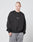A man with a shaved head models the LIVE FAST DIE YOUNG Tape Sweater in black, an oversized cotton and polyester sweatshirt, paired with light gray sweatpants against a plain light gray background.