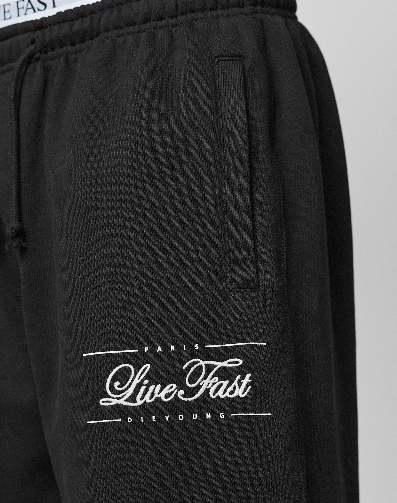 Close-up of Wide Cuff Jogger by LIVE FAST DIE YOUNG in black French Terry. Features white embroidered "LIVE FAST DIE YOUNG" text near the pocket, with visible elastic waistband and pocket opening.