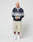 A person with short blond hair wears the LIVE FAST DIE YOUNG LFDY Striped Polo in navy and gray, beige knee-length cotton twill shorts, white socks, and white sneakers, standing against a plain light background.