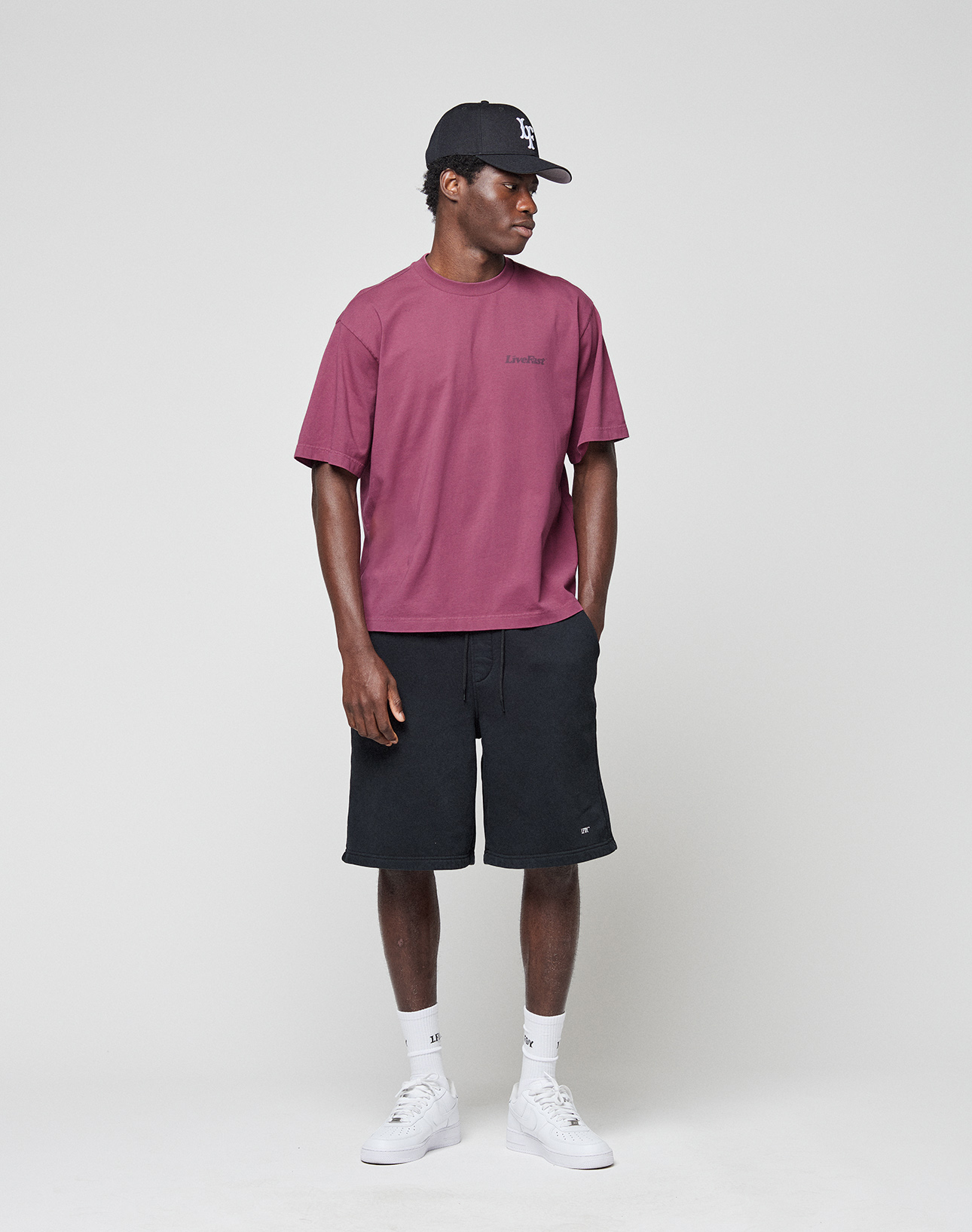 A man stands against a plain background wearing a black cap, the LIVE FAST DIE YOUNG Basic 210 Tee in maroon (100% Baumwolle), black knee-length shorts, white crew socks, and white sneakers. He looks slightly left with hands in his pockets.