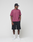 A man stands against a plain background wearing a black cap, the LIVE FAST DIE YOUNG Basic 210 Tee in maroon (100% Baumwolle), black knee-length shorts, white crew socks, and white sneakers. He looks slightly left with hands in his pockets.