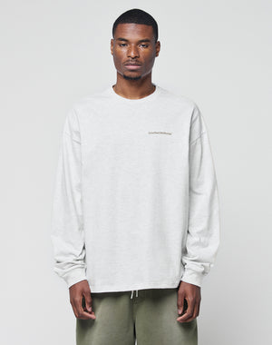 A man with short hair wears the LIVE FAST DIE YOUNG Basic Longsleeve, a light gray, logo-print, oversize sweatshirt, paired with olive green pants, standing against a plain light background.