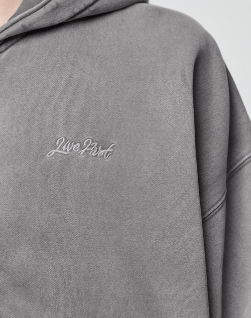 Close-up of the Basic Script Hooded Zip by LIVE FAST DIE YOUNG: a gray hoodie in soft cotton, featuring Live Fast embroidered in white cursive on the left chest and a subtly textured, vintage-inspired finish.
