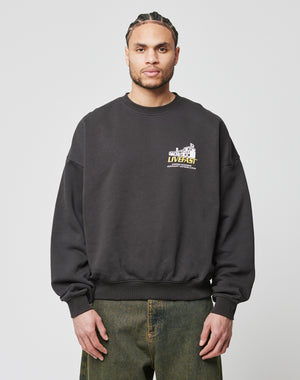 A man with braided hair wears an oversized fit black LFDY Supply Sweater by LIVE FAST DIE YOUNG, made from a Baumwolle-Polyester mix with a small cracked front print and upper left chest text, paired with greenish-grey jeans.