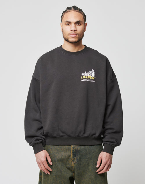 A man with braided hair wears an oversized fit black LFDY Supply Sweater by LIVE FAST DIE YOUNG, made from a Baumwolle-Polyester mix with a small cracked front print and upper left chest text, paired with greenish-grey jeans.