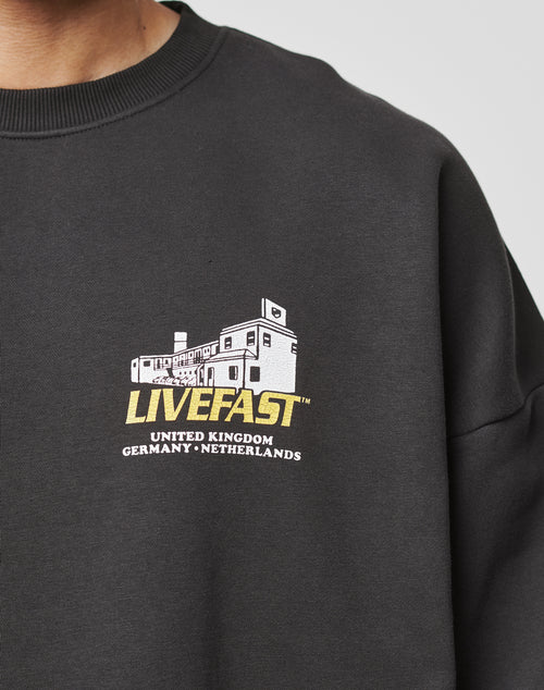 Close-up of a person in the LFDY Supply Sweater by LIVE FAST DIE YOUNG, featuring an oversized black fit with a white building graphic and cracked “LIVEFAST UNITED KINGDOM GERMANY NETHERLANDS” print. Soft Baumwolle Polyester mix.