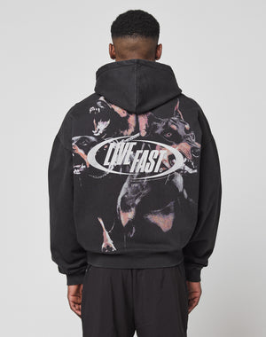 A person faces away from the camera in an oversize black LFDY Canine Hooded Zip by LIVE FAST DIE YOUNG, made from Baumwolle Polyester. It features snarling dog graphics and bold silver LIVE FAST text on the back, hood up.
