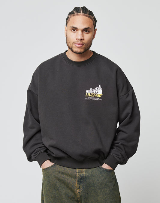 A man with braided hair wears the oversized LFDY Supply Sweater by LIVE FAST DIE YOUNG—a baggy black cotton-polyester mix with a small cracked graphic and text on the left chest—paired with loose green pants against a plain light background.