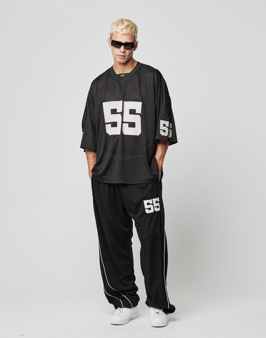 A person with short blond hair and sunglasses models LIVE FAST DIE YOUNG’s LFDY 55 Trackpants, loose black polyester track pants with an elastic waistband and white 55 detail, paired with a matching jersey and white sneakers against a plain background.