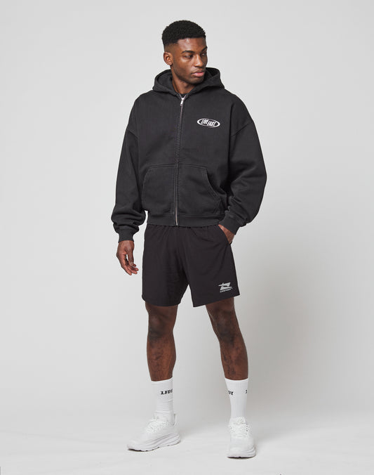 A man stands against a plain background wearing the LFDY Canine Hooded Zip by LIVE FAST DIE YOUNG, black shorts, white crew socks, and white sneakers. He has one hand in his pocket and looks to the side.