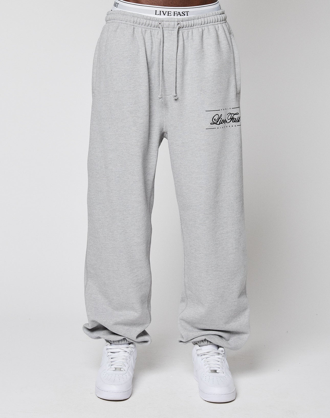 A person in light gray Wide Cuff Jogger sweatpants by LIVE FAST DIE YOUNG, featuring LIVE FAST on the waistband and thigh, plus white sneakers, stands against a plain white backdrop.