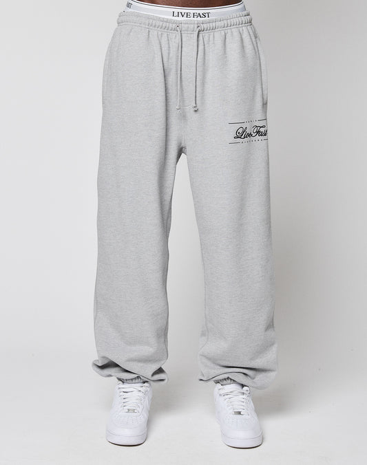 A person in light gray Wide Cuff Jogger sweatpants by LIVE FAST DIE YOUNG, featuring LIVE FAST on the waistband and thigh, plus white sneakers, stands against a plain white backdrop.