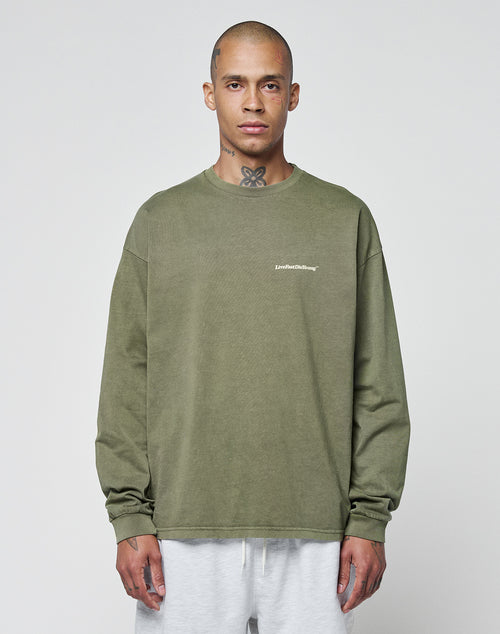 A person with a shaved head and neck tattoos wears the olive green LIVE FAST DIE YOUNG Basic Longsleeve with a white chest logo and light gray pants, standing against a plain light background.