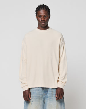 A man with braided hair wears the LIVE FAST DIE YOUNG Blank Waffle Longsleeve in cream, an oversized 100% cotton shirt, paired with light blue jeans, standing against a plain light gray background and looking at the camera.