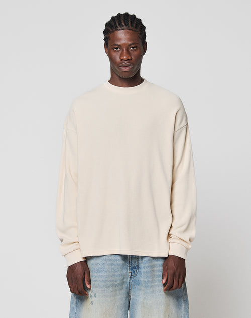 A man with braided hair wears the LIVE FAST DIE YOUNG Blank Waffle Longsleeve in cream, an oversized 100% cotton shirt, paired with light blue jeans, standing against a plain light gray background and looking at the camera.