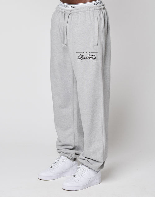 A model wears LIVE FAST DIE YOUNG Wide Cuff Jogger in light gray with "Live Fast" text on the left thigh and a comfy elastic waistband, styled with white sneakers. The photo shows the joggers waist to ankles against a plain white background.
