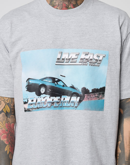 Someone wearing the LIVE FAST DIE YOUNG LFDY Crash Kid Tee—a gray cotton shirt featuring a car jumping a gap, LIVE FAST DIE YOUNG at the top, and EUROPE RUN below. The Regular Fit cut shows off their tattooed arms.