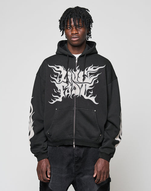 A person wears the LFDY Inferno Hooded Zip by LIVE FAST DIE YOUNG, showcasing bold white flame prints and LIVE FAME on the front with front and sleeve designs, standing against a plain light gray background.