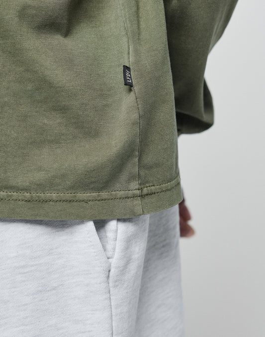 Close-up of a person wearing the LIVE FAST DIE YOUNG Basic Longsleeve in light gray (100% Baumwolle) with a pocket, olive green oversize fit, and a small black side-seam tag. Background is plain and light-colored.